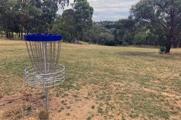 Metal disc golf catcher basket with chains sits at the top of a grassy hill with native trees to the sides and in the distance