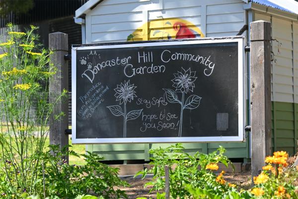 A chalkboard sign sits amongst a veggie garden with a colourful mural in the background. The sign says 'Doncaster Hill Community Garden. Stay safe. Hope to see you soon"