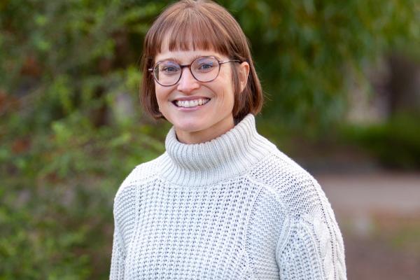 A woman stands smiling with her hands in her pockets, she has short brown hair and glasses