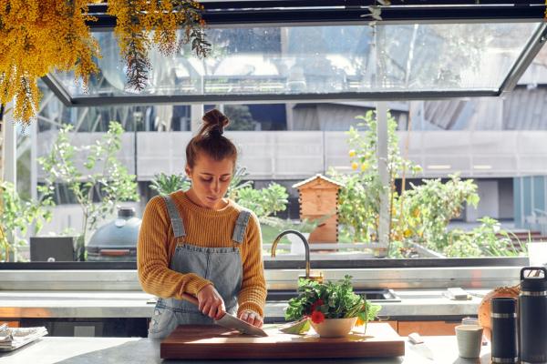 A woman chops herbs in a bright kitchen, there are fruit trees and a beehive visable out of the window