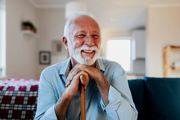 Senior man with grey hair and beard, wearing a blue button up shirt with a walking stick, sitting on a couch smiling