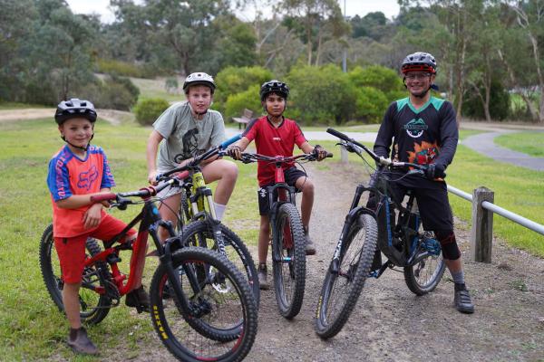 Four kids and an adult smile  to the camera, posing with their mountain bikes on a dirt path surrounded by greenery.