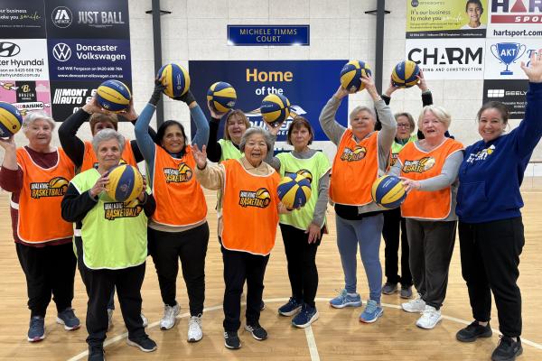 Group of 11 women standing on a basketball court each holding a basketball in the air and wearing high-vis basketball vests with their coach on the right