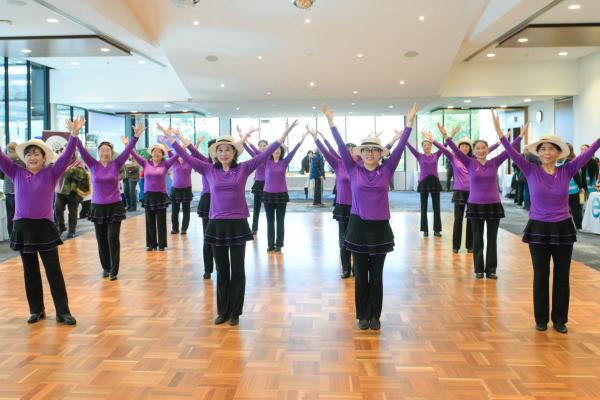A group of dancers wearing purple tops, black dressings and pants and hats, from the Seniors Happy Life group, performing at the Manningham Function Centre.