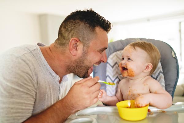 Adult male spoon-feeding a messy baby in a high chair with a yellow bowl; food is smeared on the baby's face and chest."