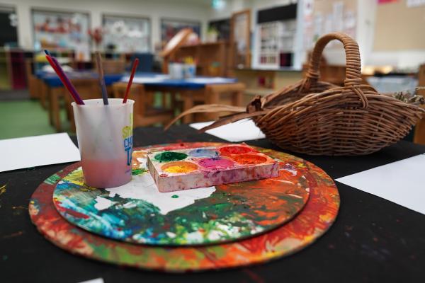 An art classroom table with a circular paint palette, paintbrushes in a cup, a tray of colourful paints, and a wicker basket. 