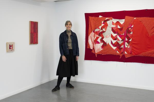 Artist Shannon Slee, wearing a black skirt and denim jacket, stands in the corner of a room next to a large red tapestry which is hanging on the while walls on the right, and a small framed red tapestry on the left.
