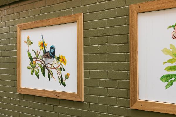 Two framed paintings on a green brick wall: one of a blue and black bird on a branch with yellow flowers, the other of green leaves and part of a plant.