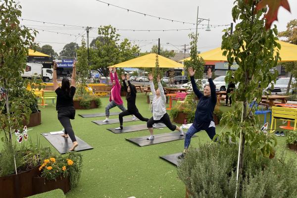 Outdoor yoga class with five people in warrior pose on black mats, surrounded by greenery and yellow umbrellas.