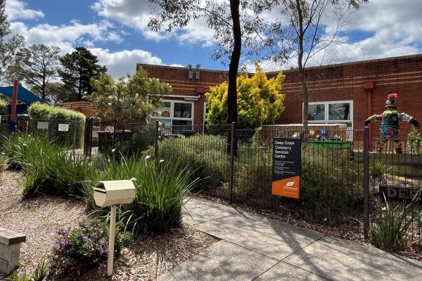 A low red brick building surrounded by a shrubby garden and a wire front fence.