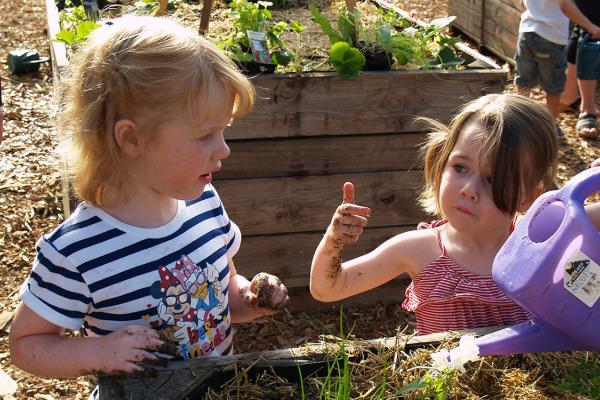 Two children gardening together in a raised garden bed. One holds a purple watering can while the other has soil on their hands. Green plants and garden beds are visible in the background.