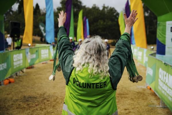 Back view of an older woman wearing a green voluneer vest, with her arms raised and holding a bunch of medallions in each hand.