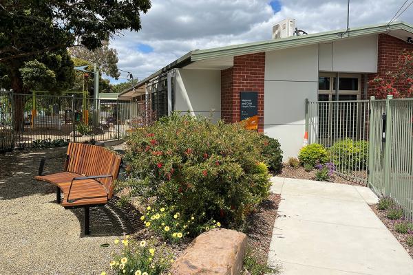 A red and white brick building surrounded by a garden with a bench set and concrete paths.