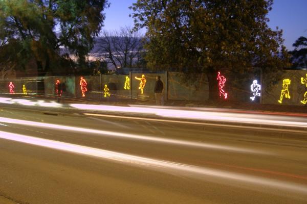 Illuminated human figures in pink, yellow and white appear on a wall alongside a road at dusk