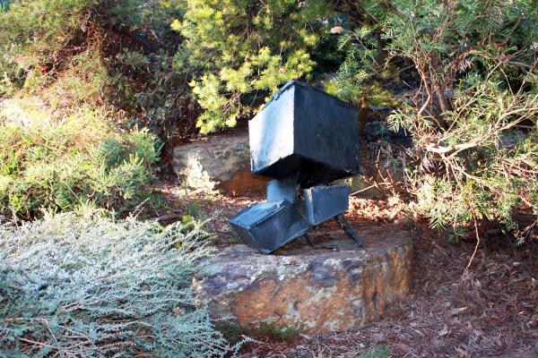 A roughly welded black steel sculpture made up of different sized cubes sits on a rocky step amongst native bushland