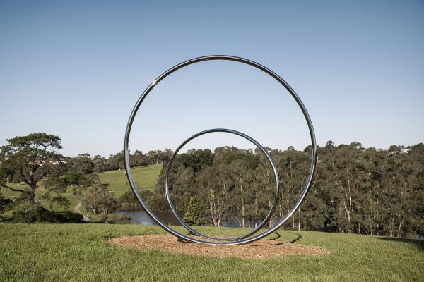 Two large polished stainless steel circles sit on a hill overlooking an open grassy expanse with gum trees and a lake in the background.