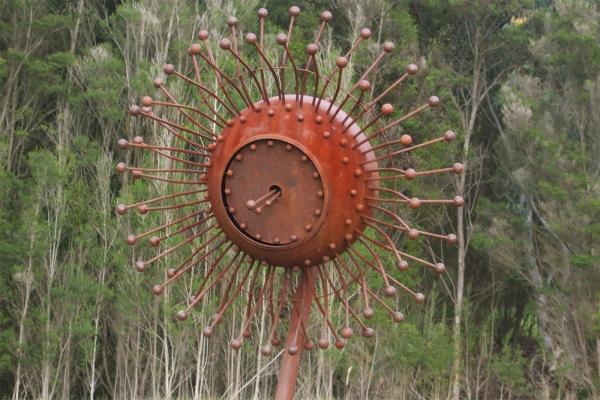 Rust coloured steel sculpture in the shape of a eucalypt blossom sits in a bushy landscape