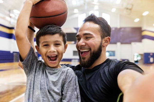 A young man taking a selfie smiling with a young boy who is holding a basketball on his head while smiling