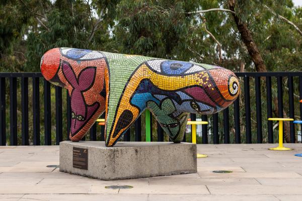 Sculpture of an abstract three-legged creature covered in small colourful mosiac tiles mounted on a concrete block, gum trees in the background.