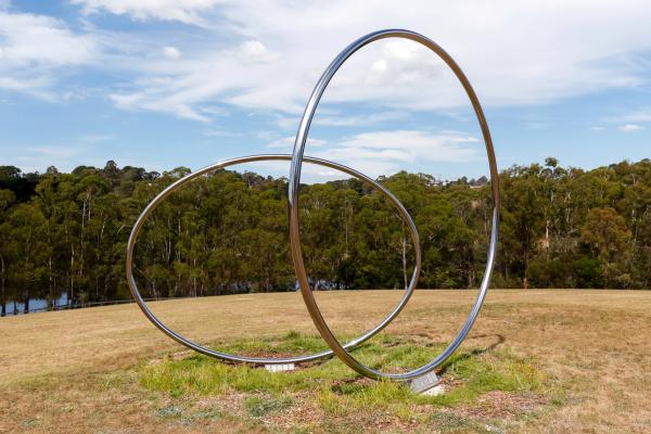 Two large polished stainless steel circles sit on a hill overlooking an open grassy expanse with gum trees and a lake in the background.