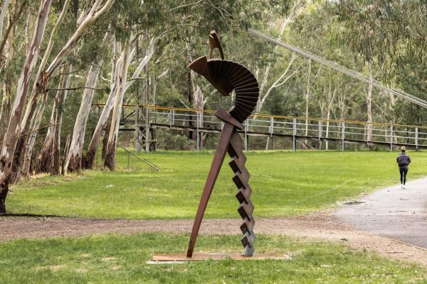 A large bronze sculpture, with a somewhat human form, stands in a bushy parkland. A bridge is in the background and a figure in black walks along a path to the left side.