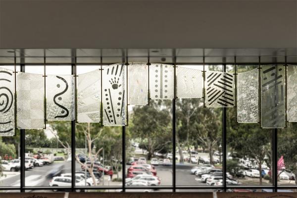 A series of 12 glass panel of different sizes, all with unique markings and textures, joined together in a wave-like pattern. A parking lot with cars and gum trees can be seen through a window behind the panels.