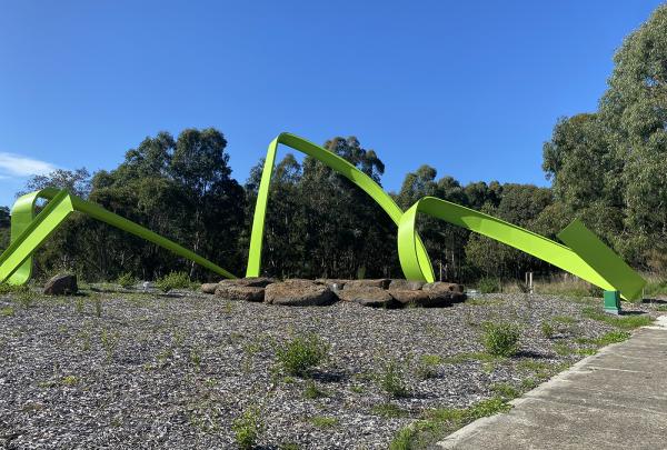 Long twisting lime green steel sculpture sits in a bushy landscape with a concrete footpath off to the right side