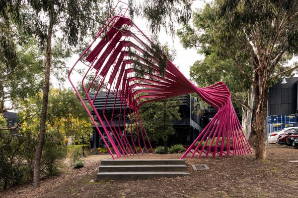 A large pink sculpture made of many bent steel poles sits in a bushy garden in front of a modern office building