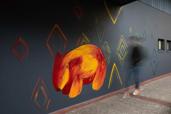 Mural of an orange and red wombat on a grey wall, a blurred figure walks in front of the wall.