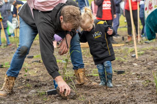 A young man and a small girl, both wearing boots, stand in a muddy field with many people behind them. The man is planting a sapling and the girl is holding his leg looking on.