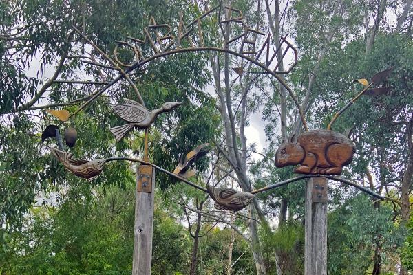 A concrete path leads up to two wooden poles with a gate between them. Above the poles sits a twisted metal sign with the words 'Wombat Bend'. A bushy parkland is in the background.