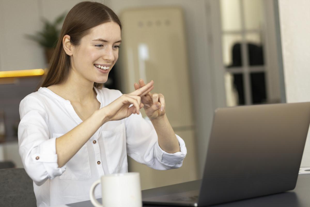 A young woman wearing a white shirt and smiling, sits in front of an open laptop doing sign language