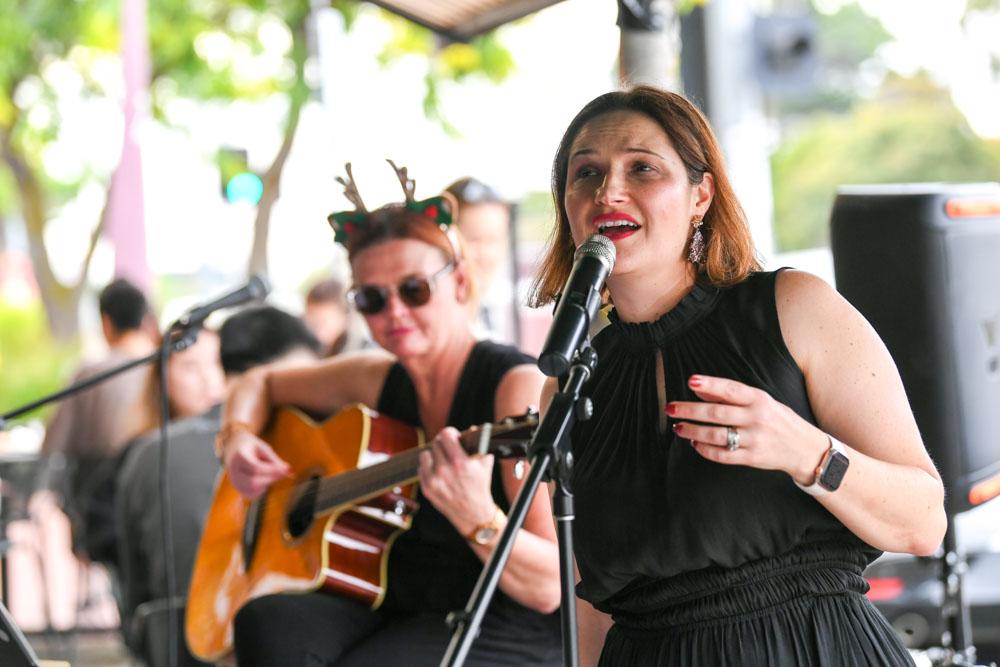 A women and a black dress singing into a microphone during an outdoor performance on the footpath with another woman wearing Christmas antler ears sitting behind her playing the guitar.