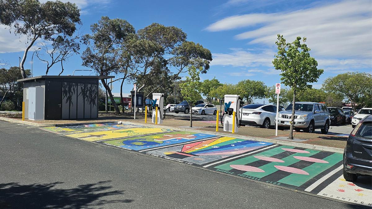 A suburban carpark with colourfully painted parking bays with EV charging stations and a toilet block to the left.