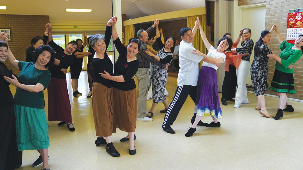 A group of older asian people dance in pairs. They are smiling and have one arm raised to the ceiling. 