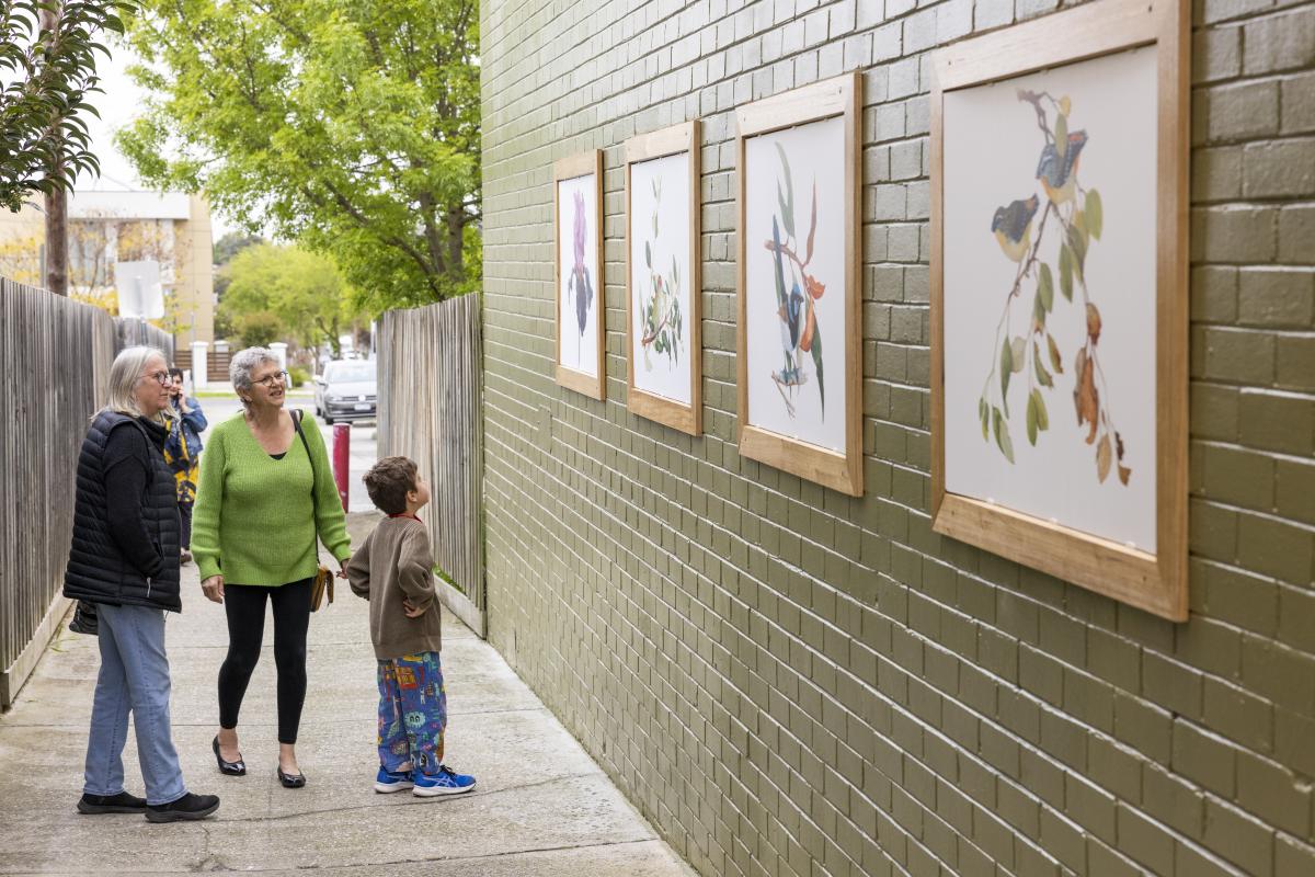 Two women and a boy looking at framed artworks by Terry Napier on the green painted brick wall of Tunstall Square laneway