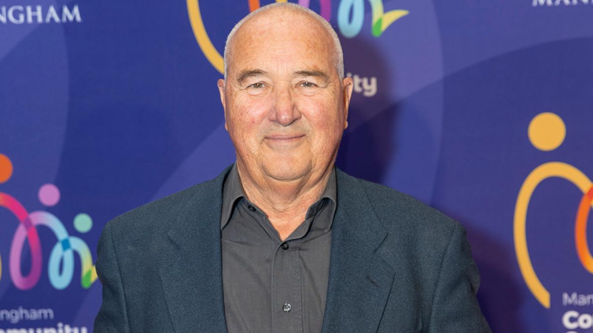 An older man in a dark coloured shirt and jacket stands smiling in front of a purple background