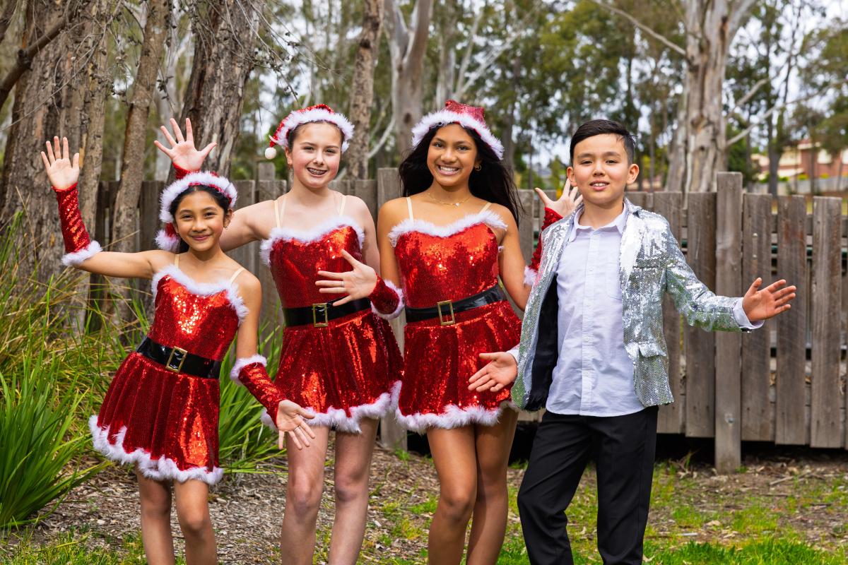 Three girls dressed in glittery red Santa style dresses with hats and handless gloves with a boy dressed in white shirt, black pants and silver glittery jacket wave to cameras.