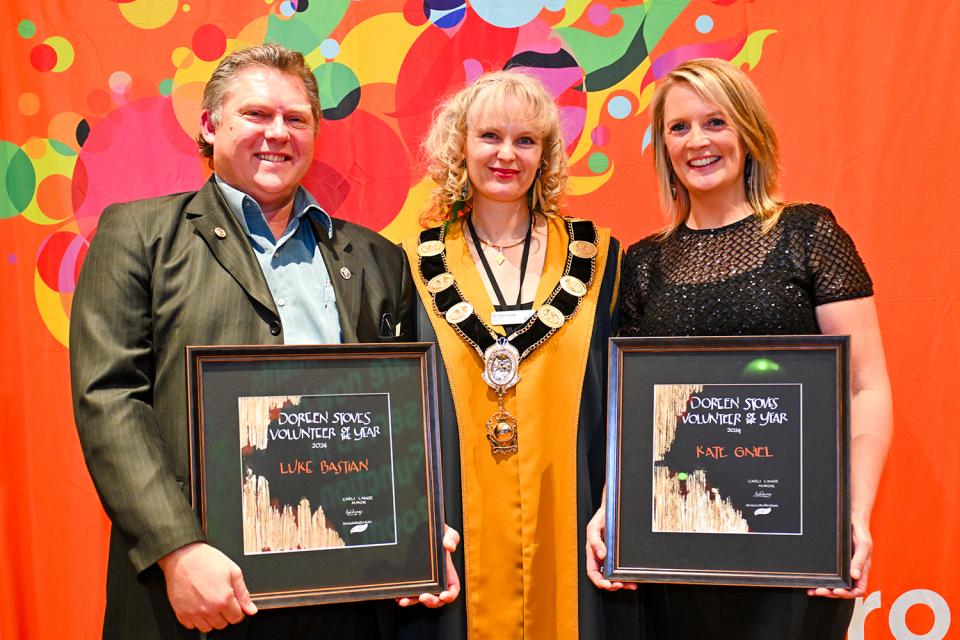 Luke Bastian, Kate Gniel and Mayor Cr Carli Lange stand smiling in front of an orange background, Kate and Luke are holding their Civic Awards.