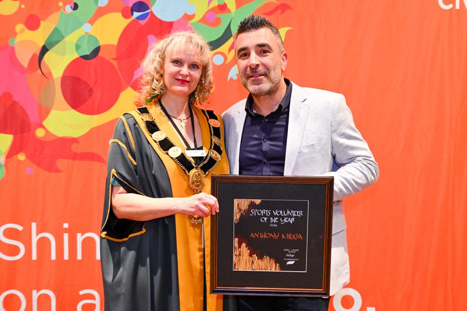 Anthony Milicia and Mayor Cr Carli Lange stand smiling in front of an orange background, Anthony is holding his Civic Award.