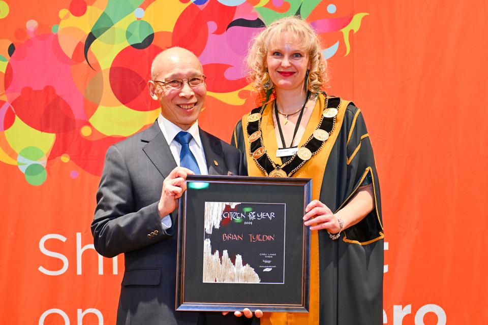 Brian Tyedin and Mayor Cr Carli Lange stand smiling in front of an orange background, Brian is holding his Civic Award.