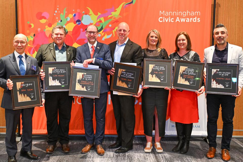 Five men and two women stand in front of an orange banner with the text Manningham Civic Awards in the top right. They are smiling and holding their awards in front of them.