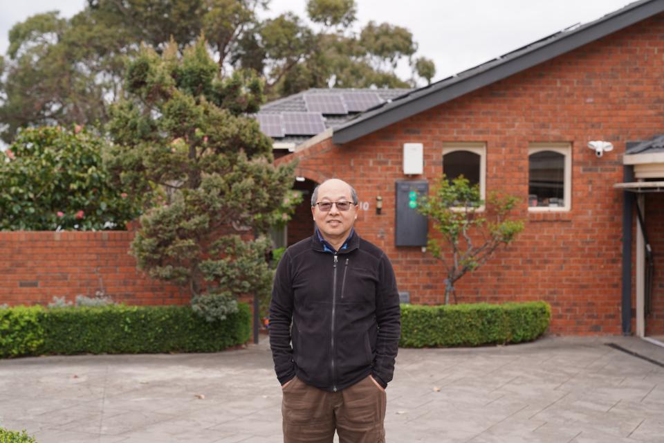 A man standing in front of a brick house with solar panels on the roof, surrounded by greenery.