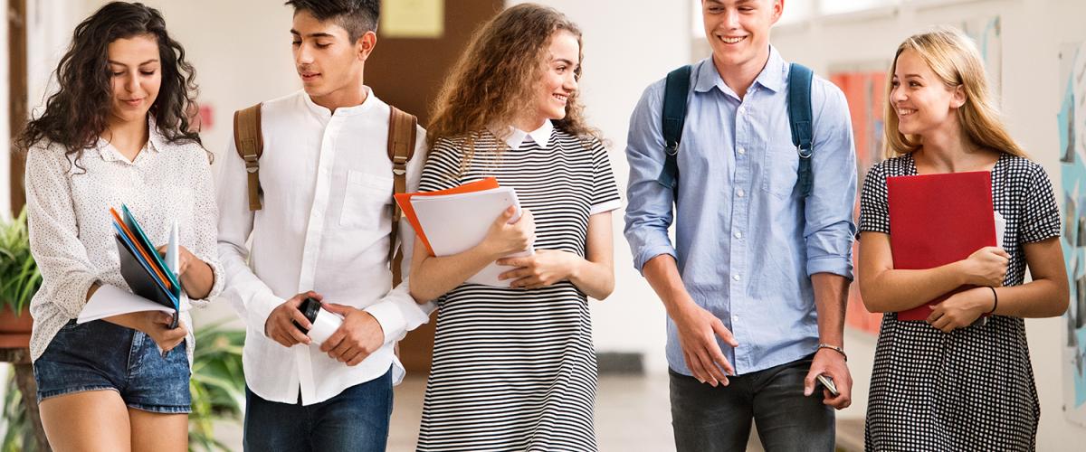 Five young adults carrying books, walking down a wide hallway, smiling and looking at each other