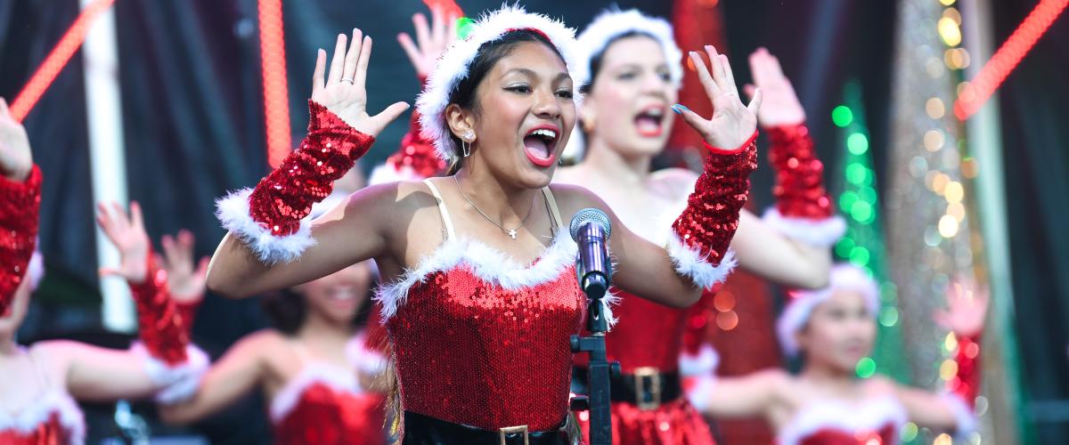 Dancers dressed in sparkly red Santa-style costumes, singing and dancing on stage at the 2024 Manningham Carols By Candlelight