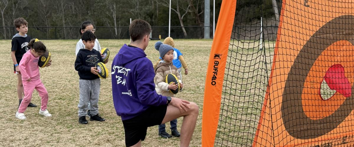 Young children lining up on an oval to throw a football through a target.