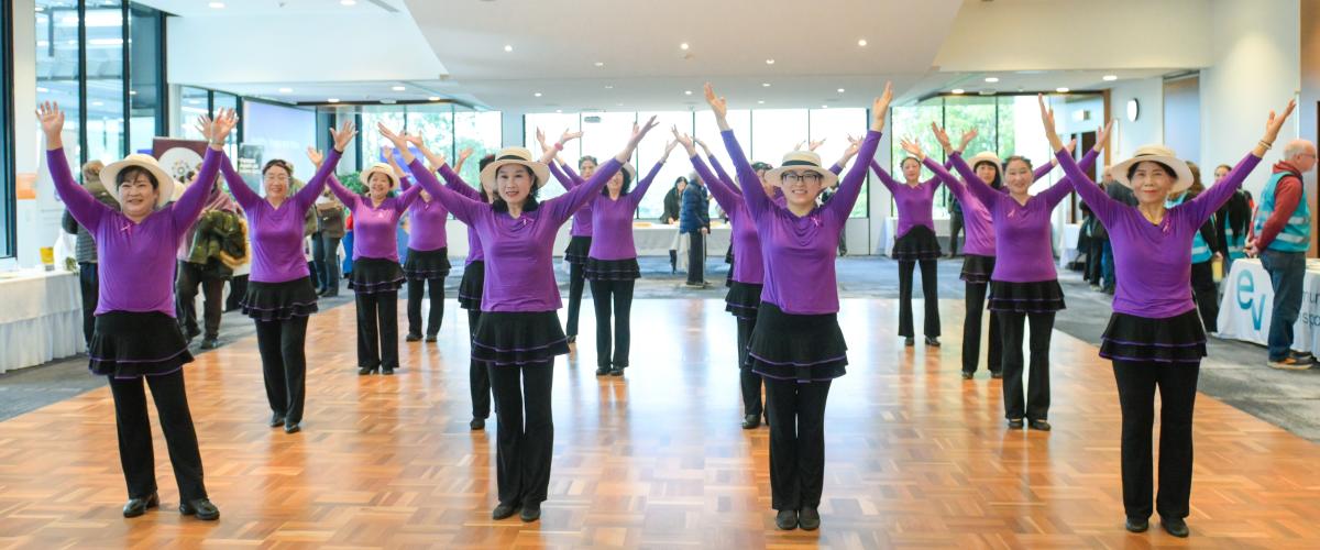 A group of dancers wearing purple tops, black dressings and pants and hats, from the Seniors Happy Life group, performing at the Manningham Function Centre.