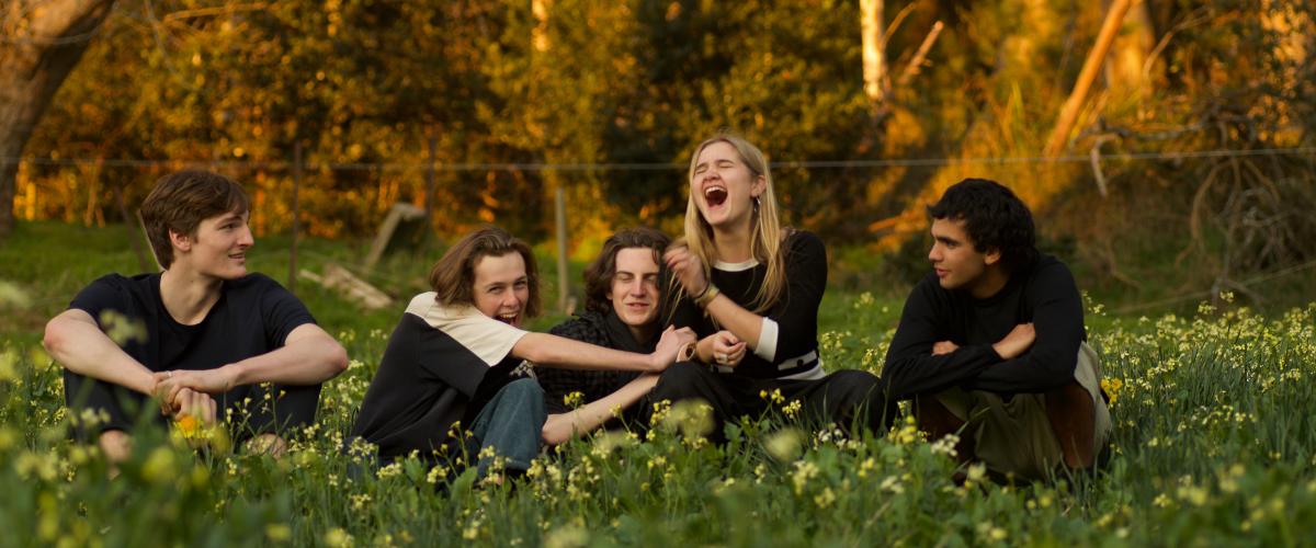 Members of the band BOTH sitting in a field of grass and yellow flowers with bushland in the background.