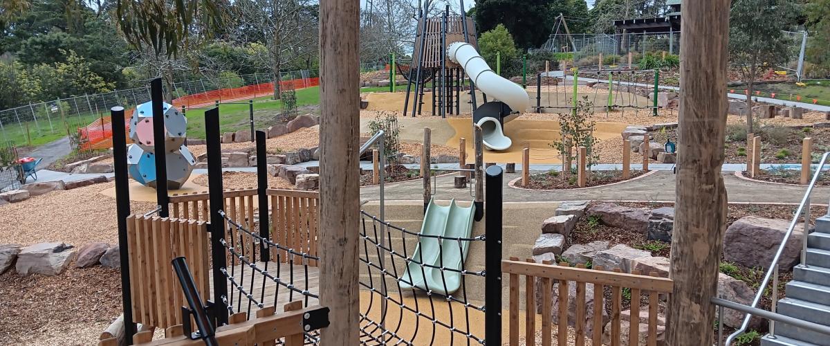 A modern playground with wooden structures with multiple slides in a bushy park setting.