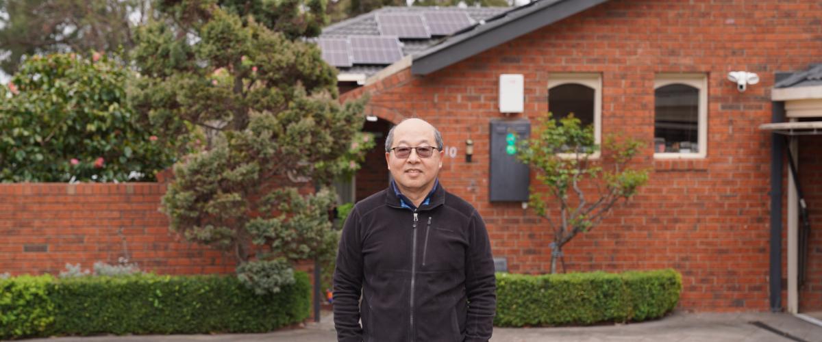 A man standing in front of a brick house with solar panels on the roof, surrounded by greenery.